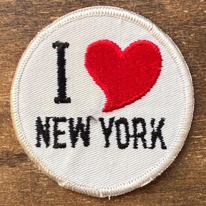 May include: A circular patch with the phrase "I ❤️ NEW YORK" embroidered in black and red on a light gray background. The heart is red, and the text is in a bold, sans-serif font. The patch has a white stitched border.