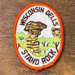 May include: Embroidered patch with an oval shape, featuring the text "Wisconsin Dells Stand Rock" in a circular pattern. The patch has a white background with a detailed illustration of a rock formation in shades of brown and yellow, surrounded by green foliage.