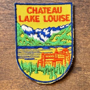 May include: Embroidered patch with the text "CHATEAU LAKE LOUISE" above a scenic design. The design features mountains, a lake, and a building, all in vibrant colors on a yellow background. The patch has a blue border.