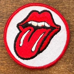 以下が含まれることがあります： ローリング・ストーンズの舌と唇のロゴをあしらった刺繍パッチ。赤い舌と唇に白い歯、黒い縁取り、白い背景に赤い縁取りのデザインです。