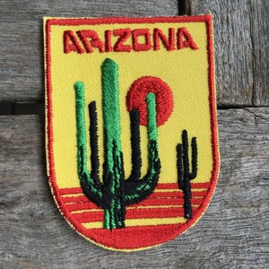May include: A yellow and red embroidered patch with the word "ARIZONA" in red lettering. The patch features a green and black cactus with a red sun behind it.