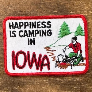 Peut inclure: Écusson brodé avec une bordure rouge et le texte "HAPPINESS IS CAMPING IN IOWA". L'écusson représente une scène de camping avec une personne cuisinant au-dessus d'un feu, deux arbres verts et une rivière. L'écusson est rectangulaire.