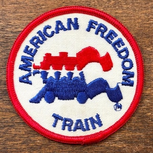 Peut inclure: Écusson circulaire avec une bordure rouge et un centre blanc. L'écusson porte les mots "AMERICAN FREEDOM TRAIN" en bleu, entourant un dessin de train bleu et rouge. L'écusson est probablement un article brodé.