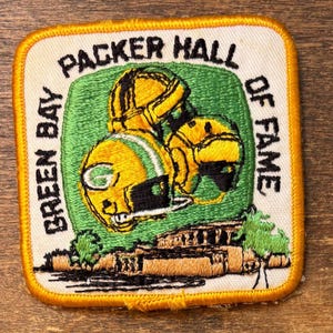 Puede incluir: Un parche cuadrado con borde amarillo presenta el texto "GREEN BAY PACKER HALL OF FAME". El parche tiene un fondo verde con dos cascos de fútbol amarillos y verdes. Un edificio marrón y verde está en la parte inferior.