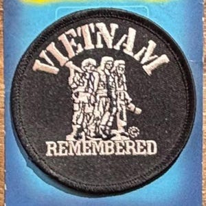 Op de afbeelding: Een ronde zwarte geborduurde patch met wit stiksel. De tekst VIETNAM staat in een boog aan de bovenkant en REMEMBERED aan de onderkant. In het midden staan drie figuren naast elkaar, afgebeeld in wit garen op de zwarte achtergrond.