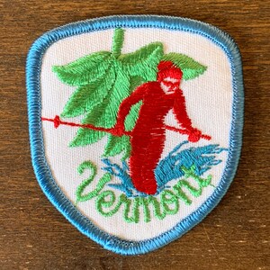 以下が含まれることがあります： 青い縁取りのある白い刺繍パッチ。パッチには、緑の木の中に赤いスキーヤーが描かれており、緑色の刺繍で「Zornig」という単語が書かれています。