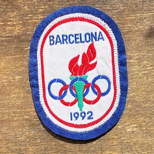 May include: An oval-shaped embroidered patch featuring a white background with a blue border. The text BARCELONA is at the top and 1992 is at the bottom. In the center, a green torch with a red flame is positioned over the five Olympic rings.