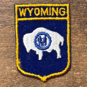 May include: A navy blue patch with a gold border and the word "WYOMING" at the top. The patch features a white bison with a blue and white emblem in the center. The patch is a shield shape.