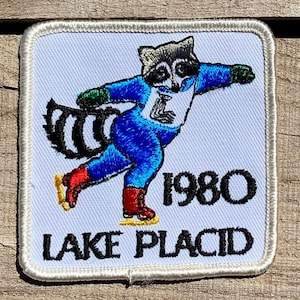 Peut inclure: Un écusson brodé blanc représentant un raton laveur faisant du patin à glace. Le raton laveur porte une combinaison de patinage bleue et blanche avec un bonnet rouge et blanc. L'écusson porte l'inscription "1980 Lake Placid" en lettres noires.