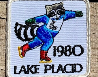 Puede incluir: Un parche blanco bordado con un mapache patinando sobre hielo. El mapache lleva un traje de patinaje azul y blanco con un gorro rojo y blanco. El parche dice "1980 Lake Placid" en letras negras.
