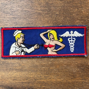 May include: A rectangular embroidered patch with a blue background and red border. It features a cartoon sailor in a white uniform using a stethoscope on a blonde figure wearing a red bikini top, alongside a white caduceus medical symbol.