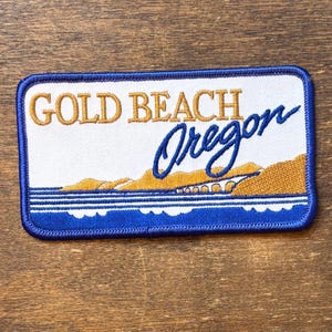 Könnte beinhalten: Ein rechteckiger bestickter Aufnäher mit dem Text GOLD BEACH in Goldfaden und Oregon in blauer Schreibschrift. Das Design zeigt eine goldene Küstenlandschaft mit einer Brücke über blauem Wasser, eingefasst von einem dicken blauen Rand.