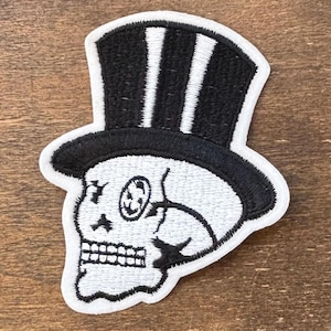 May include: Embroidered patch featuring a skull wearing a black top hat with white stripes. The skull is white with black details, including the eye and mouth. The patch has a white border.