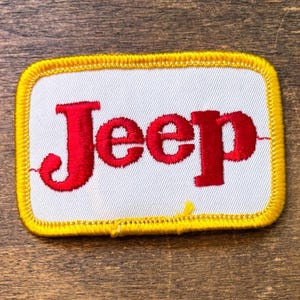 May include: Embroidered patch featuring the word "Jeep" in red, centered on a white rectangular background. The patch has a yellow border with a stitched design. The patch is likely for clothing or accessories.