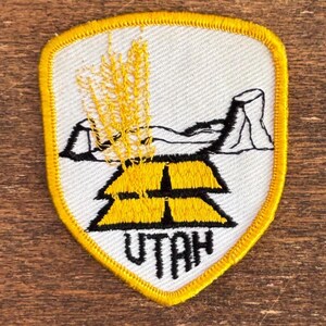 May include: Embroidered patch featuring a shield shape with a yellow border. The patch depicts a stylized landscape with yellow wheat, a rock formation, and the word "UTAH" in black lettering. The background is white.