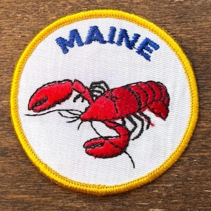 May include: Embroidered patch featuring a red lobster on a white background. The word "MAINE" is embroidered in blue above the lobster. The patch has a yellow border.