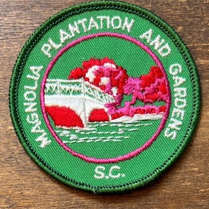 May include: A round green patch with the words "Magnolia Plantation and Gardens" in white text. The patch features a bridge and trees in red, white, and pink, with "S.C." at the bottom. The patch has a pink inner circle.