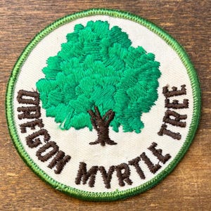 Könnte beinhalten: Ein runder, bestickter Aufnäher mit einem leuchtend grünen Baum mit braunem Stamm auf cremefarbenem Hintergrund. Der Text OREGON MYRTLE TREE ist in braunem Garn entlang der unteren Kurve gestickt und von einem grünen Rand umgeben.