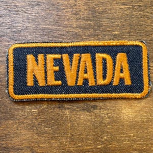 May include: A rectangular embroidered patch featuring the word NEVADA in bold, golden-yellow thread. The lettering is set against a dark navy blue background, and the entire patch is bordered by a thick, golden-yellow stitched edge.