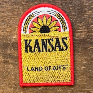 Puede incluir: Parche bordado con borde rojo, con la palabra "KANSAS" en letras negras sobre un fondo dorado. El parche incluye un diseño de girasol y el texto "LAND OF AH'S".