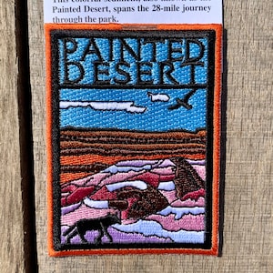Puede incluir: Parche bordado con las palabras "PAINTED DESERT" sobre un fondo de cielo azul. El parche representa un paisaje desértico con formaciones rocosas coloridas, un animal negro y un pájaro. El parche tiene un borde naranja.