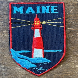 May include: A shield-shaped embroidered patch with a dark blue background and a red border. It features the word MAINE in light blue letters above a red and white lighthouse. Yellow lines represent light beams, and light blue waves are at the base.