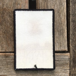 Op de afbeelding: Een witte patch met zwarte stiksels rond de randen. De patch is leeg en heeft een klein zwart touwtje dat eronder hangt.