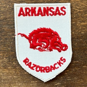 Pode incluir: Um emblema bordado em forma de escudo com fundo branco e borda vermelha. A palavra ARKANSAS está bordada em vermelho no topo, com um gráfico de um javali Razorback vermelho no centro e a palavra RAZORBACKS bordada em vermelho abaixo.