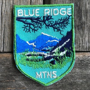May include: Embroidered patch featuring a mountain landscape with the text "BLUE RIDGE MTNS." The patch has a teal background with blue mountains, green grass, and a dark blue tree branch. The border is green.