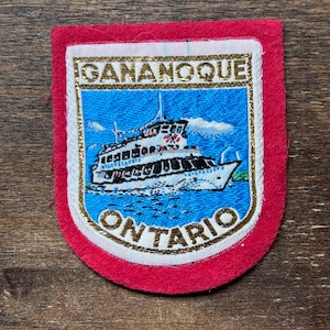 May include: A red, white and gold embroidered patch with the words "GANANOQUE ONTARIO" and a depiction of a white ferry boat on blue water.