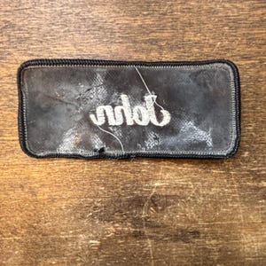以下が含まれることがあります： 白い刺繍文字が入った長方形の黒いパッチ。パッチには黒い縁取りがあり、木製の表面に配置されています。テキストは「nddb」と読めるようです。