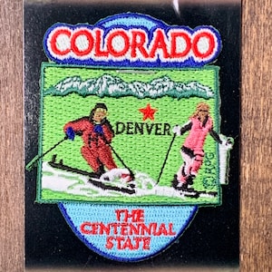 May include: A blue and red embroidered patch with the words "Colorado, The Centennial State" and a depiction of skiers in a snowy mountain landscape. The patch also includes the words "Denver" and a red star.