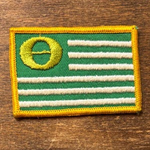 Könnte beinhalten: Gestickter Aufnäher mit grünem Hintergrund, weißen horizontalen Streifen und einem gelben Emblem. Der rechteckige Aufnäher hat einen gelben Rand. Das Design erinnert an eine Flagge.
