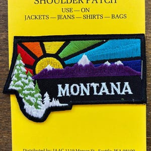May include: A black and white embroidered patch with a rainbow mountain scene and the word "MONTANA" in white text. The patch is designed to be sewn onto jackets, jeans, shirts, or bags.