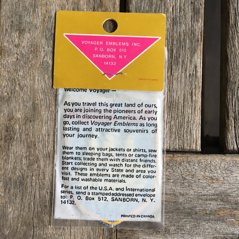 May include: A Voyager Emblems Inc. patch in clear plastic packaging with a gold header card featuring a pink triangle. The card displays the company name, address, and text encouraging collectors to wear or sew the emblems on various items.