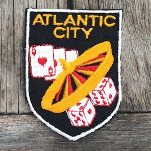 May include: Black embroidered patch with the words "ATLANTIC CITY" in gold. The patch features a casino theme with playing cards, a roulette wheel, and dice. The patch has a white border.
