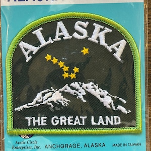 May include: Green and white embroidered patch with the text "ALASKA THE GREAT LAND" and a yellow star constellation. The patch is surrounded by a green border.