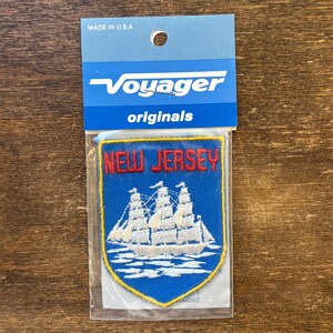 May include: A shield-shaped embroidered patch featuring a white sailing ship on a blue background with the red text NEW JERSEY above it. The patch is enclosed in a clear plastic sleeve with a blue header card labelled Voyager originals.