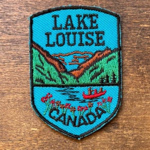 May include: Embroidered patch featuring a scenic view of Lake Louise in Canada. The design includes a lake with a red canoe, mountains, and the text "LAKE LOUISE CANADA" in black lettering against a turquoise background.