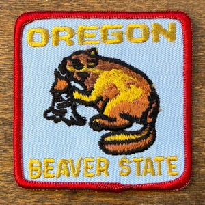 Peut inclure: Écusson brodé représentant un castor brun et jaune tenant une branche d'arbre. L'écusson a un fond bleu clair avec les mots "OREGON" et "BEAVER STATE" en lettres dorées, encadrés d'une bordure rouge.