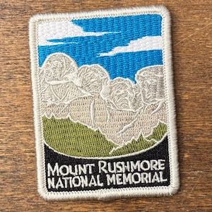 May include: Embroidered rectangular patch depicting Mount Rushmore National Memorial. The design features the presidents' faces, a blue sky with white clouds, green grass, and the text "Mount Rushmore National Memorial" in white.