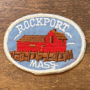 Pode incluir: Um emblema bordado oval com fundo azul claro que mostra um edifício vermelho sobre um cais de madeira castanho. O texto ROCKPORT está arqueado a branco sobre o edifício e a palavra MASS está bordada a branco sob o cais.