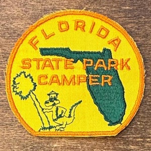 May include: A circular embroidered patch with a yellow background and an orange border. It features a green silhouette of the state of Florida, a green palm tree, and a green alligator wearing a hat. The text reads FLORIDA STATE PARK CAMPER.