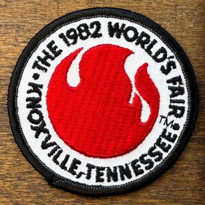 May include: Circular patch with a black border and white background. The patch features the text "Knoxville, Tennessee" and "The 1982 World's Fair" in black. A red flame design is in the center of the patch.