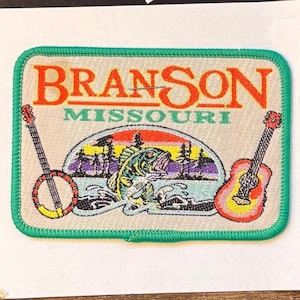 May include: A rectangular patch with the words "BRANSON MISSOURI" in orange and green. The patch features a colorful illustration of a fish jumping out of water, with a banjo and guitar on either side. The patch has a green border.