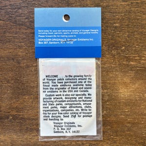 May include: A rectangular clear plastic packaging sleeve with a blue header card featuring a circular hole. The card contains printed text regarding Voyager Originals, Voyager Emblems Inc., and a mailing address in Sanborn, N.Y. 14132.