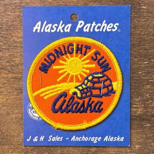 May include: A circular embroidered patch on a blue backing card labeled Alaska Patches. The patch features an orange background with a yellow sun, a blue igloo, and the blue text Midnight Sun Alaska. The card includes the text J & H Sales - Anchorage Alaska.