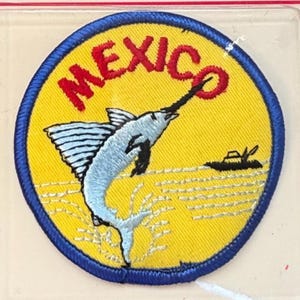 May include: Circular embroidered patch with a yellow background and a blue border. It features a light blue marlin leaping from the water, with the word "MEXICO" in red above. A small boat is also depicted.