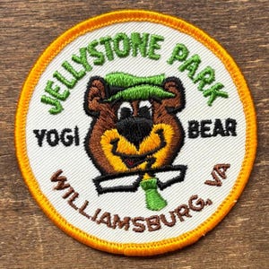 May include: Embroidered patch featuring Yogi Bear from Jellystone Park in Williamsburg, VA. The patch has a white background with a yellow border. The bear is brown with a green hat and tie, and black glasses. The text "Yogi Bear" and "Jellystone Park" are in green.