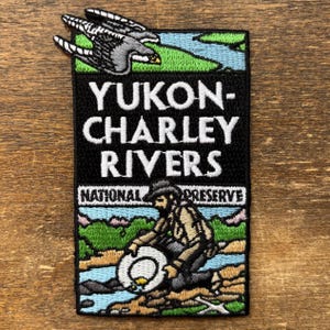 May include: Embroidered patch with the text "YUKON-CHARLEY RIVERS NATIONAL PRESERVE." The patch is a black rectangle with an illustration of a bird and a person panning for gold. The design includes blue, green, and brown colours.